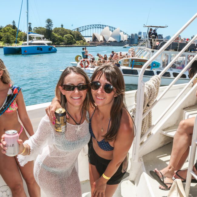 A group of people in swimwear enjoys a sunny day on a boat. Two women in the foreground pose with drinks and sunglasses. The backdrop features calm water, trees, and Sydney Opera House.