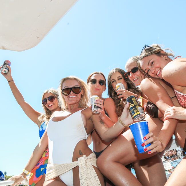 A group of people in swimwear enjoying a sunny day on a boat. They are smiling, holding drinks, and posing closely together against a bright blue sky.