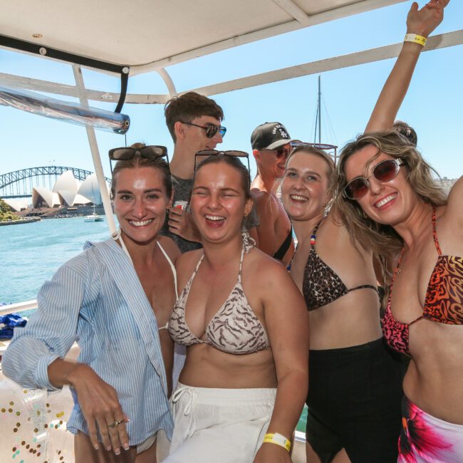 A group of people in swimwear smiling and posing on a boat in sunny weather. The background features the Sydney Opera House and Sydney Harbour Bridge, with more people on boats enjoying the day.