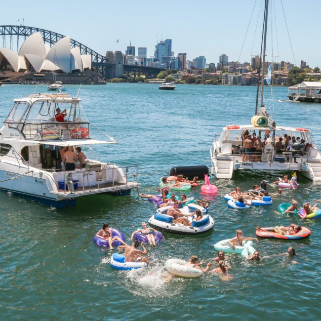 A lively scene on a sunny day featuring two boats on the water with numerous people floating nearby on colorful inflatables. In the background, the Sydney Opera House and Sydney Harbour Bridge are visible against the city skyline.