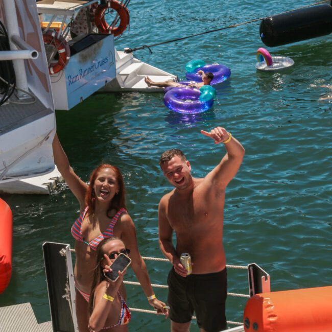 People on a boat enjoy a sunny day, with one person holding a drink and two others posing playfully near the water. Inflatable floats are visible in the water nearby. The scene is festive and lively.