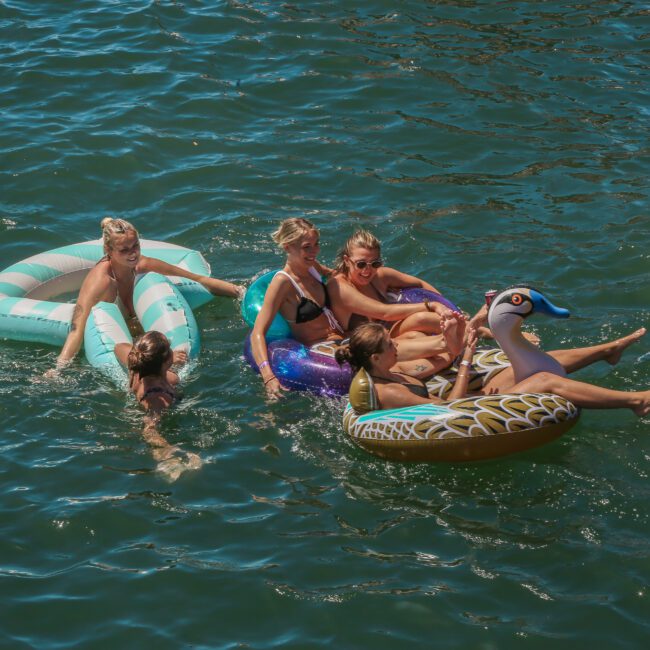 A group of people relaxing on inflatable pool floats on a lake. They are enjoying the sunny weather and are close together, with a mix of different float designs, including a bird-shaped float. The water is a deep green-blue.