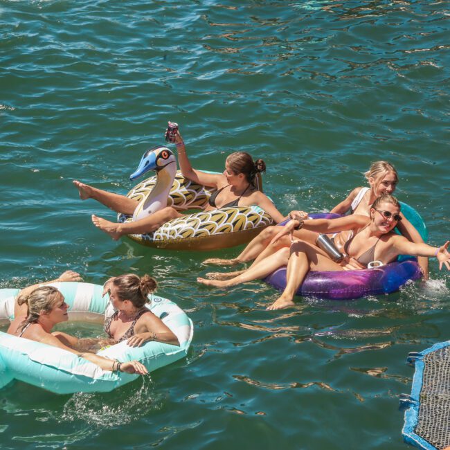 A group of people relax on inflatable floats in a sunny river. They appear to be laughing and enjoying the water, with some floats shaped like animals. The water is a deep greenish-blue, reflecting the clear, sunny sky.