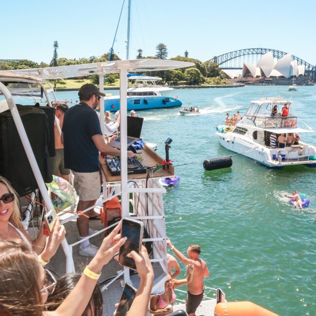 People enjoying a lively boat party on a sunny day, with a DJ playing music. Boats and people relaxing on inflatables are on the water. The Sydney Opera House and Sydney Harbour Bridge are visible in the background.