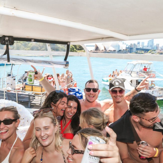 A group of people enjoying a lively party on a boat under sunny skies. They are laughing, dancing, and holding drinks, with scenic views of the water and distant city skyline in the background. Other boats are visible nearby.