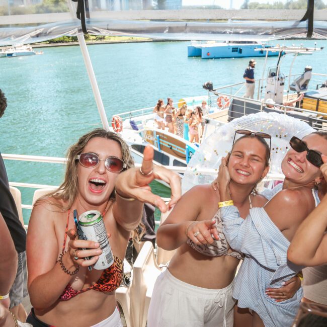 A lively group of people partying on a boat in swimwear. They're smiling, holding drinks, and enjoying the sunny day. Other boats and people are visible on the water in the background.