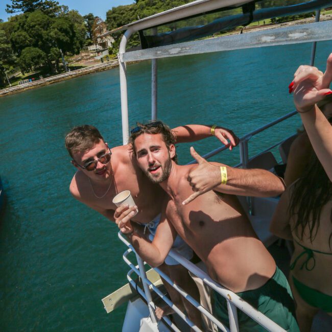 Two men on a boat enjoying a sunny day. One is holding a drink and giving a thumbs-up, while the other is smiling. Both are shirtless. The backdrop features a calm body of water and lush greenery.