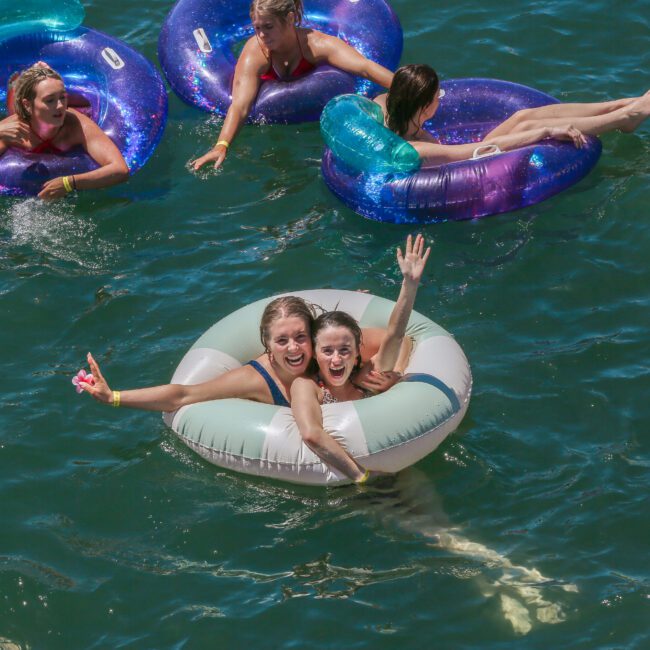 A group of people enjoying a sunny day in a lake, floating on inflatable rings. Two smiling individuals wave excitedly at the front, while others relax in the background on blue and yellow tubes. The water is clear and vibrant.