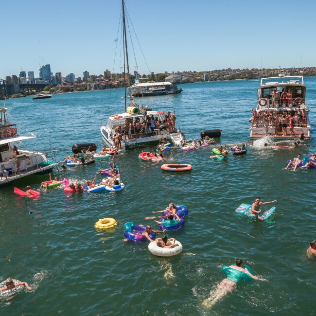 People enjoying a sunny day in a bay, swimming and floating on inflatables between several boats. Sydney Opera House and the city skyline are visible in the background under a clear blue sky.