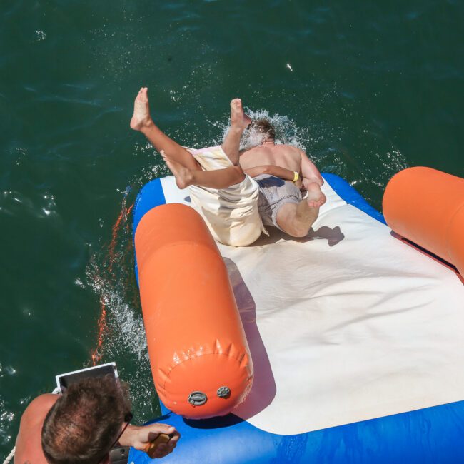 A person is playfully sliding off an inflatable water trampoline and splashing into the water. The trampoline has bright orange and blue sections. Another person is sitting nearby, enjoying the sunny day at the water's edge.