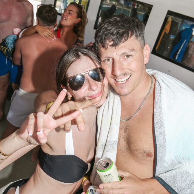 A man and woman pose playfully on a boat. The woman is wearing sunglasses and making a peace sign, while the man holds a drink and has a towel over his shoulder. Other people are in the background, some embracing and smiling.