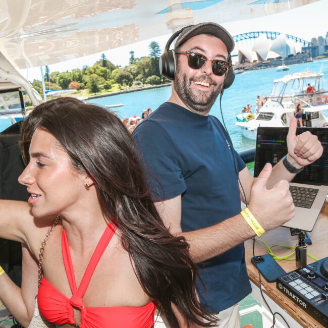 A DJ wearing sunglasses and headphones gives a thumbs-up next to a woman in a red top, against a backdrop of vibrant waterfront party scenes with boats and buildings in the distance.