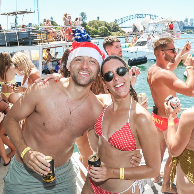 A group of people in swimsuits enjoying a sunny day on a boat party. A man in a Santa hat and a woman in a red bikini smile at the camera. Other partygoers in the background hold drinks, with the Sydney Harbour Bridge visible in the distance.