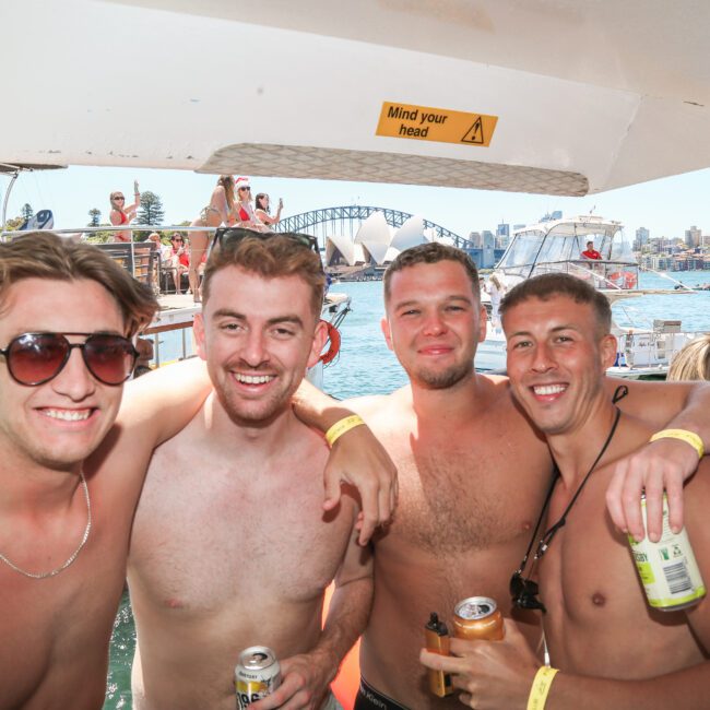 Four shirtless men are smiling and posing together on a boat. They are holding drinks, and other people and boats are visible in the background on a sunny day. A "Mind your head" sign is above them, with a cityscape and bridge in the distance.