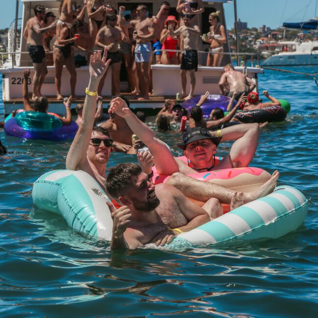 A group of people enjoying a sunny day on the water, with several individuals lounging on inflatable floats. Others are gathered on a boat in the background, with a cityscape visible beyond. The scene is lively and festive.