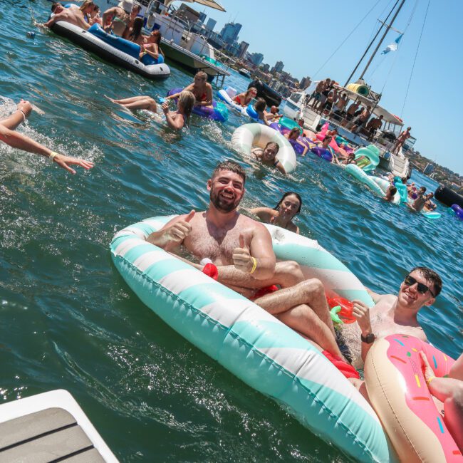 A group of people enjoying a sunny day on the water, lounging on colorful inflatable rafts. Others swim nearby, with a backdrop of boats and a distant view of a city skyline. The atmosphere is festive and relaxed.