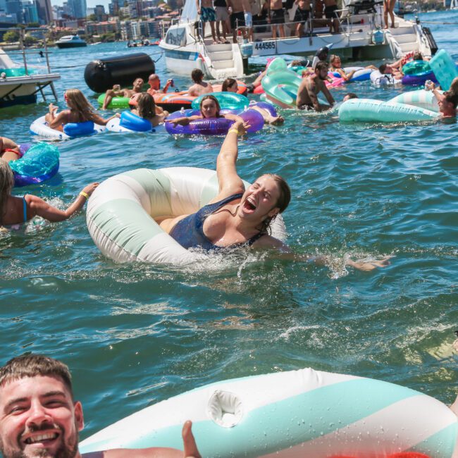 A group of people enjoying a sunny day on the water, floating on inflatable rings. Smiling faces and laughter abound as people relax and splash around. Boats and a city skyline are visible in the background.