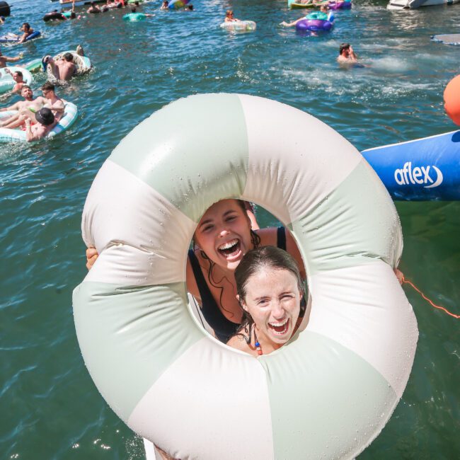 Two people smiling through a large inflatable ring on a sunny day at a lake. The water is filled with swimming people and boats in the background.