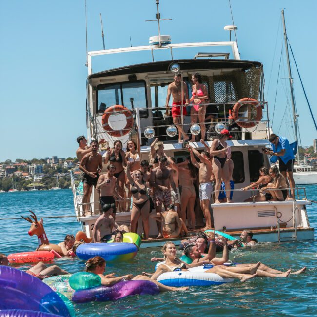 A group of people enjoying a sunny day on a boat and in the water, surrounded by colorful inflatable pool toys. The background shows a clear blue sky and distant shoreline.