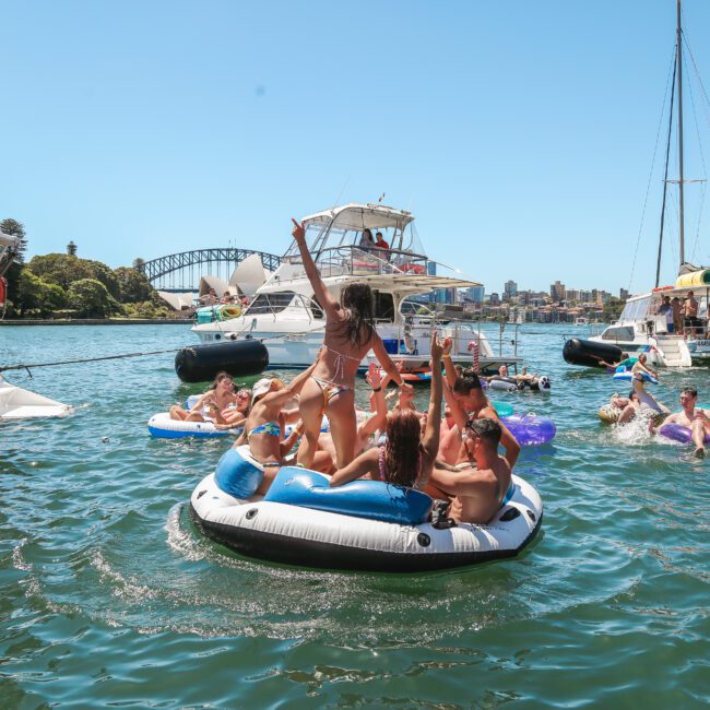 A group of people on an inflatable raft in the water enjoying a sunny day. Other boats and people are nearby, with a city skyline and bridge in the background. Some are floating on unicorn inflatables.