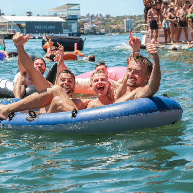 A group of men joyfully float on an inflatable raft in a sunny, crowded harbor. They are surrounded by people swimming and relaxing on boats. The water sparkles under the bright sunlight. They are smiling and appear to be having fun.