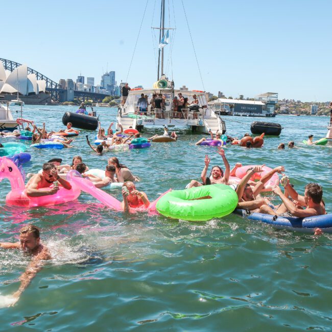 A lively scene of people floating on inflatable pool toys in a sunny harbor. Boats are anchored nearby, and the city skyline with a bridge is visible in the background. Some people are swimming and enjoying the water.