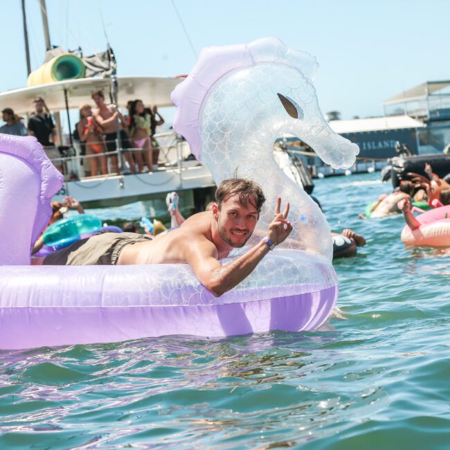 A man lounges on a large inflatable seahorse in a body of water, smiling and making a peace sign. Others float nearby on inflatables under a sunny sky, with a boat and buildings in the background.