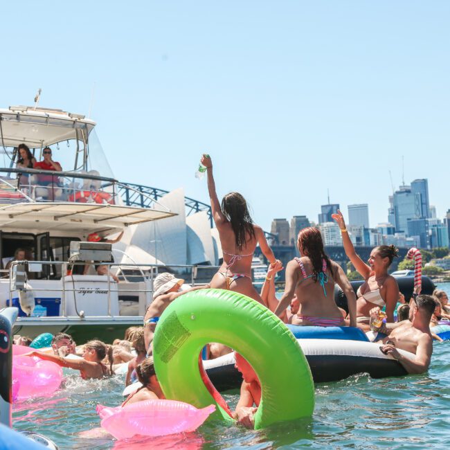 People enjoying a sunny day on the water with inflatable pool toys. A boat is docked nearby, and a city skyline is visible in the background. The scene is lively and festive.