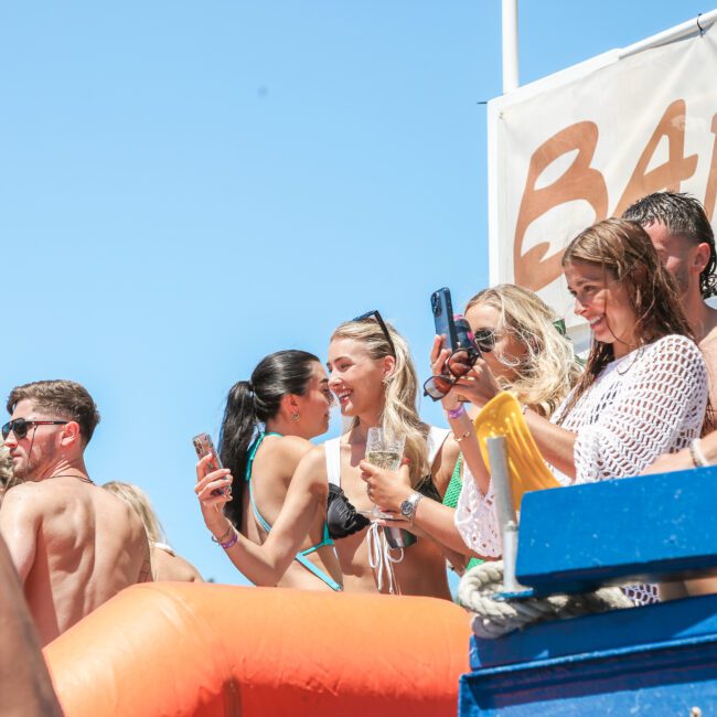 A group of people on a boat deck enjoying a sunny day. They are wearing swimwear, holding drinks, and using their phones. Some are capturing the moment with photos. A blue sky is in the background.