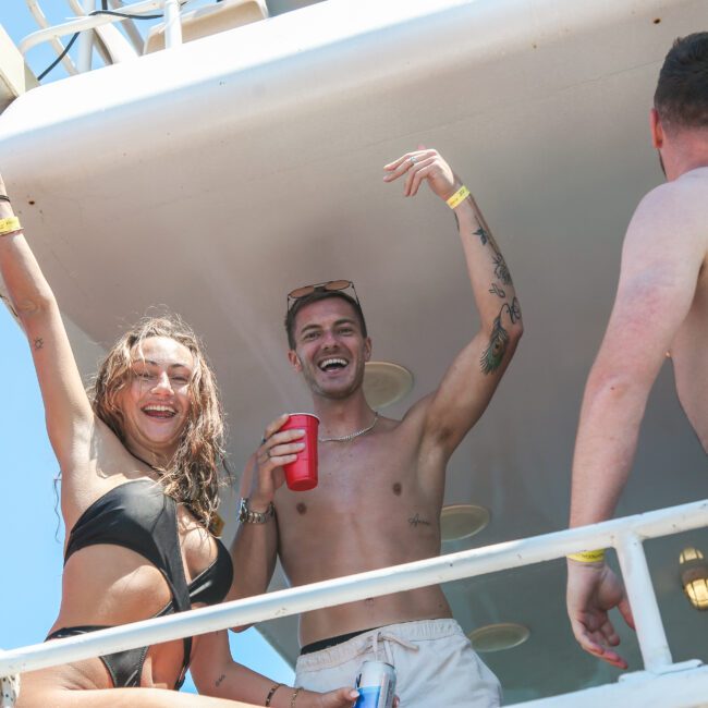 Two people in swimsuits smile and raise their arms on a boat, with drinks in hand. Another person, wearing shorts, stands nearby. The sky is clear and the mood appears celebratory.