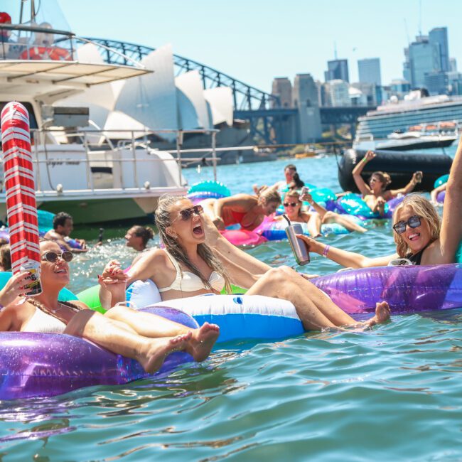 People are enjoying a sunny day on colorful inflatables in the water. They're laughing and raising their hands, with boats and a city skyline in the background.