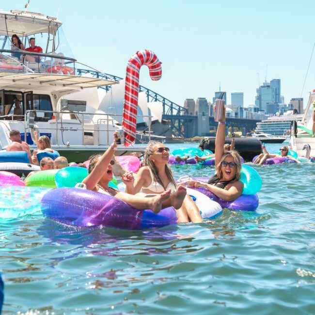 People are enjoying a sunny day on the water, lounging on inflatable floats. Boats are anchored nearby, and a large inflatable candy cane decoration is visible. The city skyline can be seen in the background.