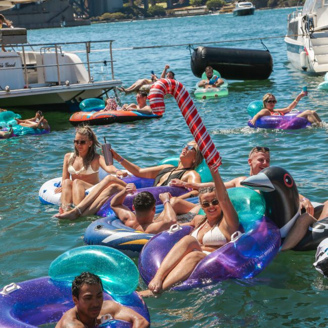 A group of people relax on inflatable floats in a sunny harbor. Some hold drinks, and one person waves a large inflatable candy cane. Boats and a city skyline are visible in the background.