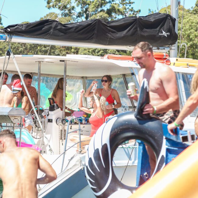 A group of people enjoying a sunny day on a boat. Some are lounging on inflatable swans, while others are socializing and taking photos. The boat is surrounded by clear water, with trees visible in the background.