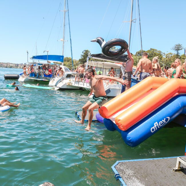 A man jumps into the water from an inflatable slide on a boat, surrounded by people on boats and floats in a sunny, festive lake setting.