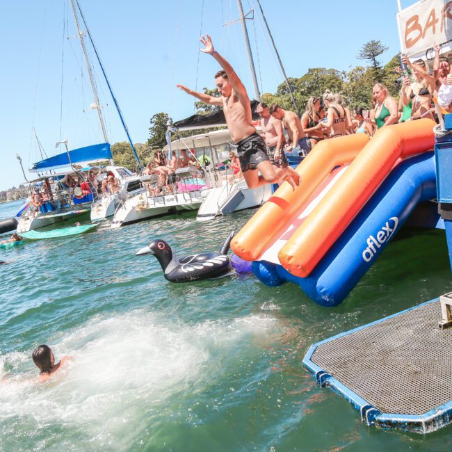 A person jumps off an inflatable slide into the water at a lively outdoor event. People watch from boats and the dock. Bright, sunny day with clear skies.