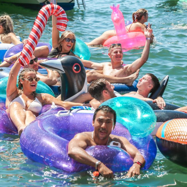 A group of people enjoying a sunny day on a lake, lounging on colorful inflatable floats shaped like animals and candy. The water is sparkling, and everyone looks cheerful and relaxed.