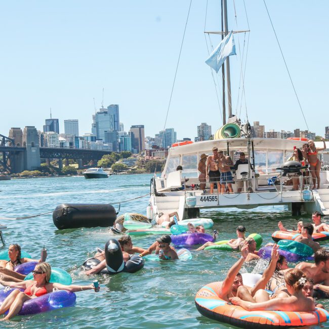 A group of people float on inflatable rings in a sunny harbor, with a boat anchored nearby. The Sydney Opera House and Harbour Bridge are visible in the background under a clear blue sky.