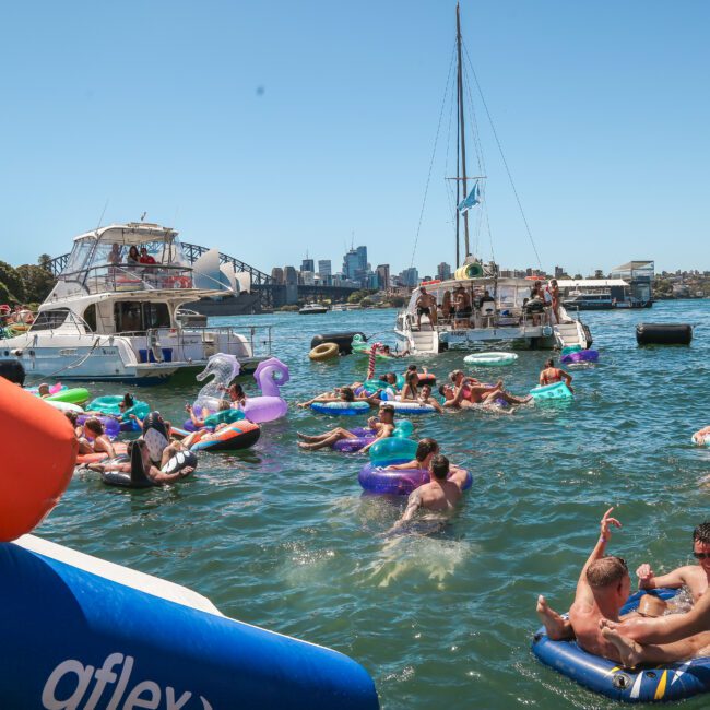 People enjoying a sunny day on a river or bay, surrounded by boats and floats. Many are swimming or relaxing on inflatable devices. The city skyline is visible in the background, under a clear blue sky.