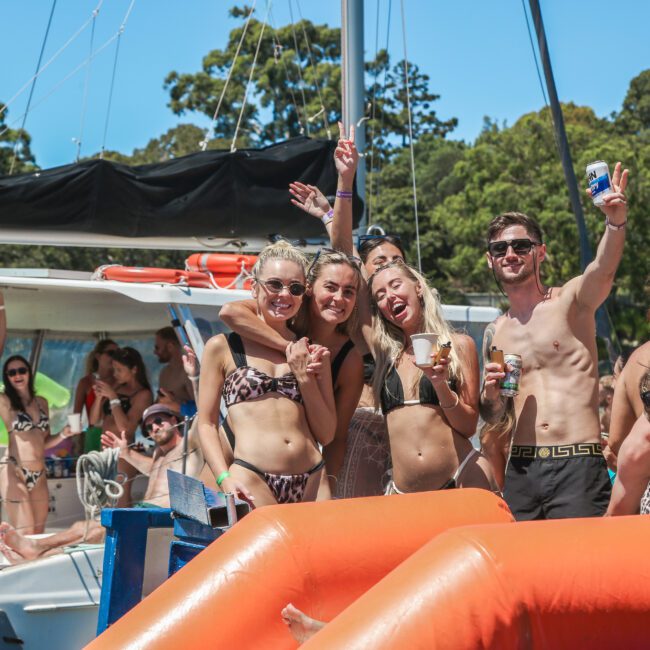 A group of people enjoying a sunny day on a boat, surrounded by trees. They are wearing swimwear, some holding drinks, and smiling at the camera. The scene is lively and festive, suggesting a summer celebration or outing.