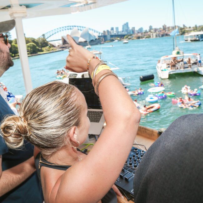 DJ on a boat plays music for a crowd on floats in a bay. A bridge and city skyline appear in the background. Bright sunny day with clear blue skies.