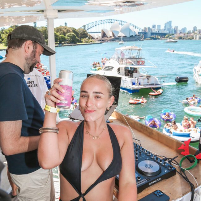 A woman in a black swimsuit raises a can while smiling on a boat with DJ equipment. A man stands nearby. Behind them, people on boats and inflatables enjoy a sunny day on the water. The Sydney Opera House and Harbour Bridge are visible in the background.