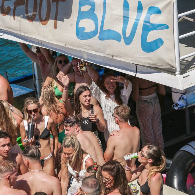 A crowded party scene on a boat with people dancing and socializing. Many hold drinks and wear swimwear. A large sign reads "BLUE" above the crowd. The atmosphere is lively and sunny, indicating a fun outdoor event on the water.