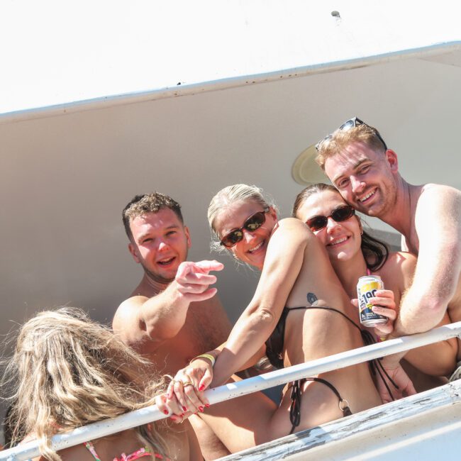 A group of people in swimwear enjoying a sunny day on a boat. Two women and two men are smiling, with one man pointing toward the camera. The scene conveys a joyful and relaxed atmosphere.