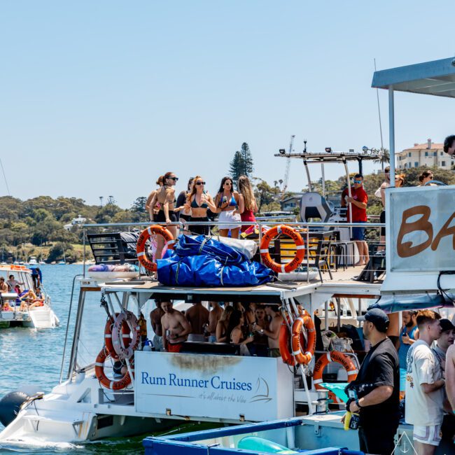 A lively boat party on a sunny day with people dancing and socializing on the upper deck. The boat, labeled "Rum Runner Cruises," is adorned with life rings and blue tarps. Other boats and scenic greenery are visible in the background.