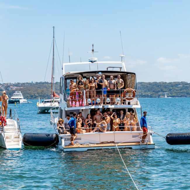 A group of people are having a party on a boat in a sunny bay. Several boats are anchored close together, with people enjoying themselves on decks. The background shows a hilly shoreline and a clear blue sky.