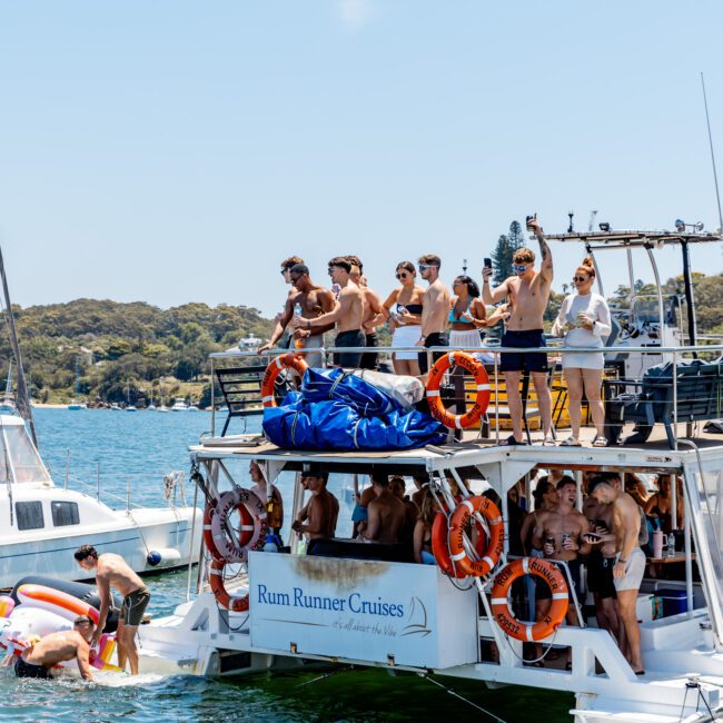 People are enjoying a sunny day on a double-deck boat named "Rum Runner Cruises." Some are sunbathing and waving on the upper deck, while others swim and relax in the water nearby. Life rings are visible on the boat's side.