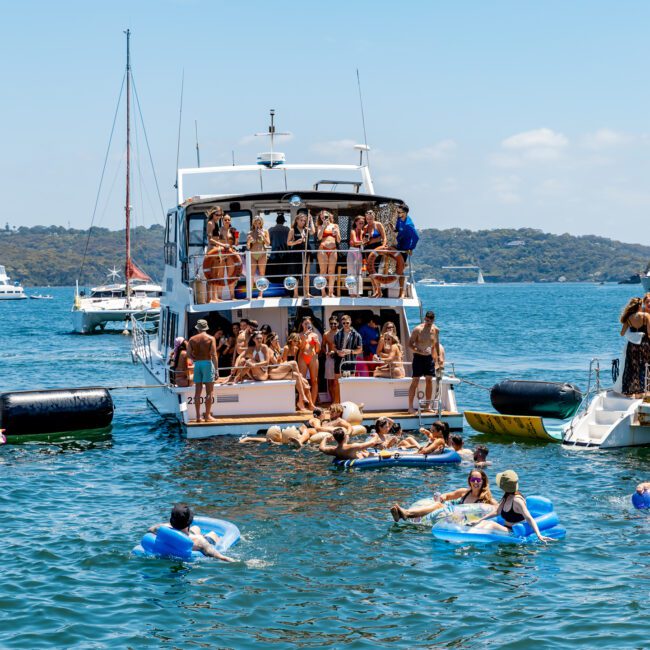 A lively gathering on a yacht and surrounding boats anchored in clear blue waters. People are swimming, lounging on inflatables, and socializing under a sunny sky. Several other boats are visible in the background with verdant hills on the horizon.