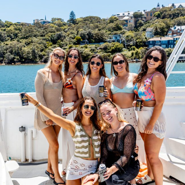 A group of seven women smiling and holding drinks on a boat. They are wearing summer outfits, with a scenic backdrop of water and green trees. The day appears sunny and bright.