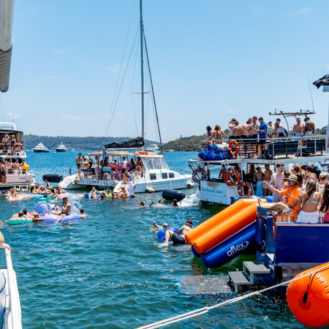 A lively scene on the water with several boats and many people enjoying a sunny day. Groups are seen relaxing, swimming, and socializing on inflatables and boats in a vibrant blue body of water.